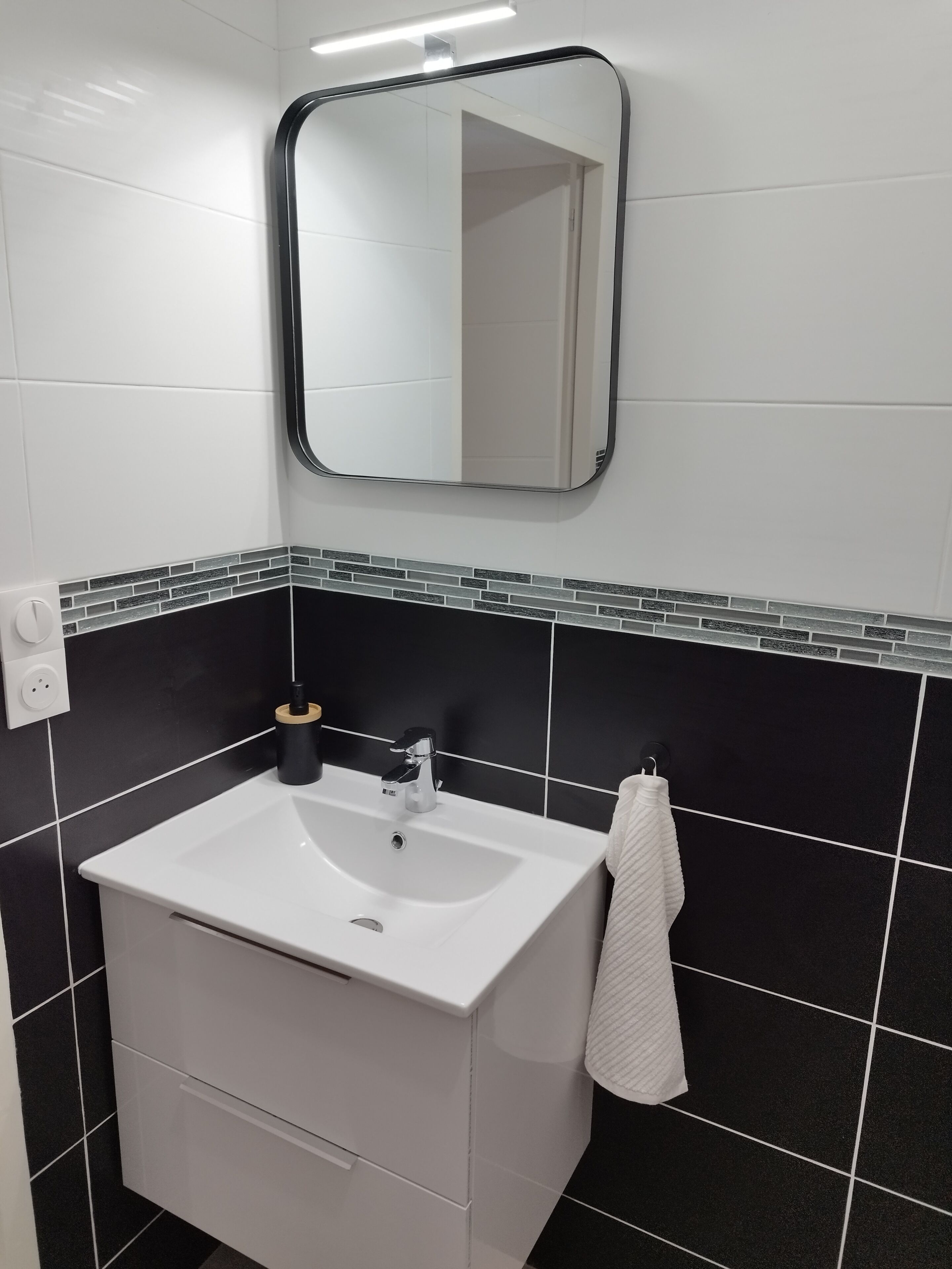 Une salle de bain avec un lavabo blanc, des murs recouverts de carreaux noirs, un miroir et une serviette accrochée au mur.