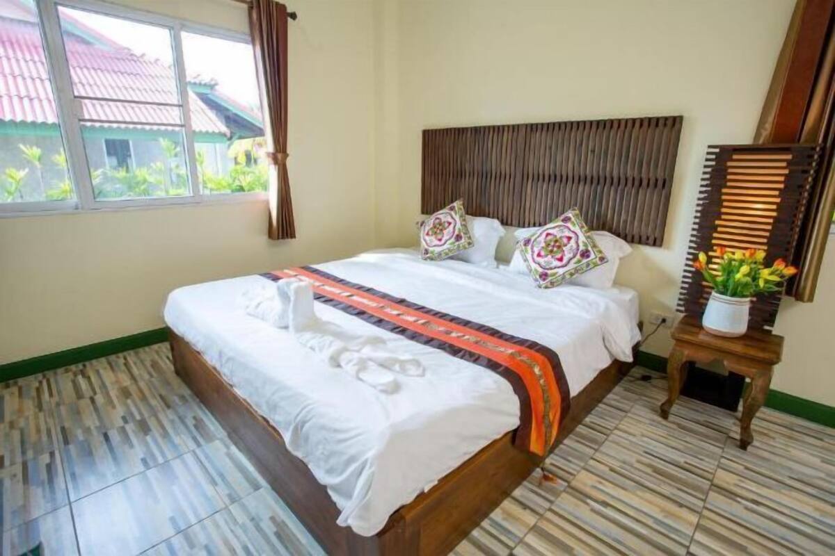 Ein Hotelzimmer mit Bett, Holz-Kopfteil, Fenster mit Vorhängen, kleinem Tisch mit Pflanze und Fliesenboden.