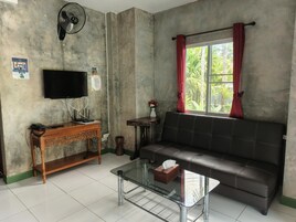 Uma sala de estar com sofá, mesa de centro de vidro, televisão, console de madeira e ventilador.