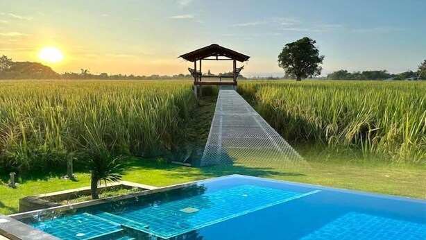 A swimming pool with a view of a rice field and a gazebo structure in the background. - Phanumas LongStay Resort  (San Pa Thong)