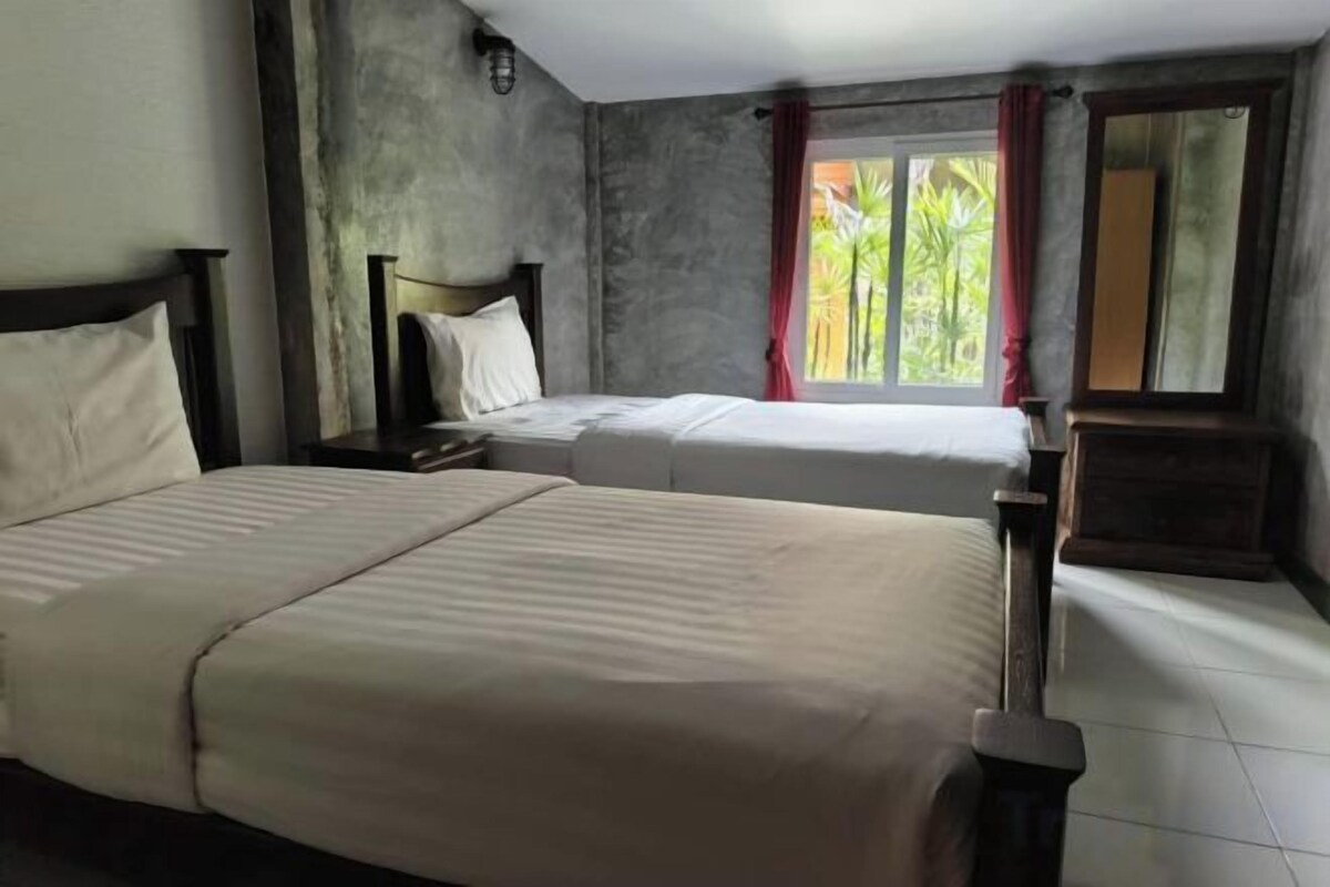 A hotel room with two beds, a window with curtains, and a view of greenery outside.