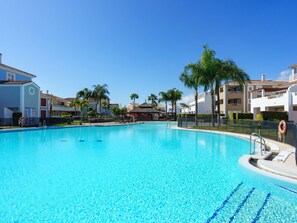 Ein klarer, blauer Pool, umgeben von weißen Gebäuden mit Balkonen, Palmen und wolkenlosem Himmel.