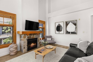 A living room with a stone fireplace, a flat-screen TV mounted above it, a comfortable sofa, and two framed artworks on the wall. - Stunning Capitol Peak Condo! Steps to Dining & Trails in Base Village.  Hot Tubs, A/C, W/D, Parking (Snowmass Village)