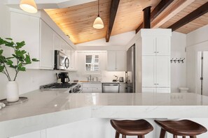 A modern kitchen with white cabinetry, a marble island, wooden ceiling beams, and a large window.