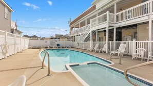 A pool area with a clear blue pool, white lounge chairs, and a two-story building with balconies. - Modern 2 Bedroom Condo with Pool (Wildwood Crest)