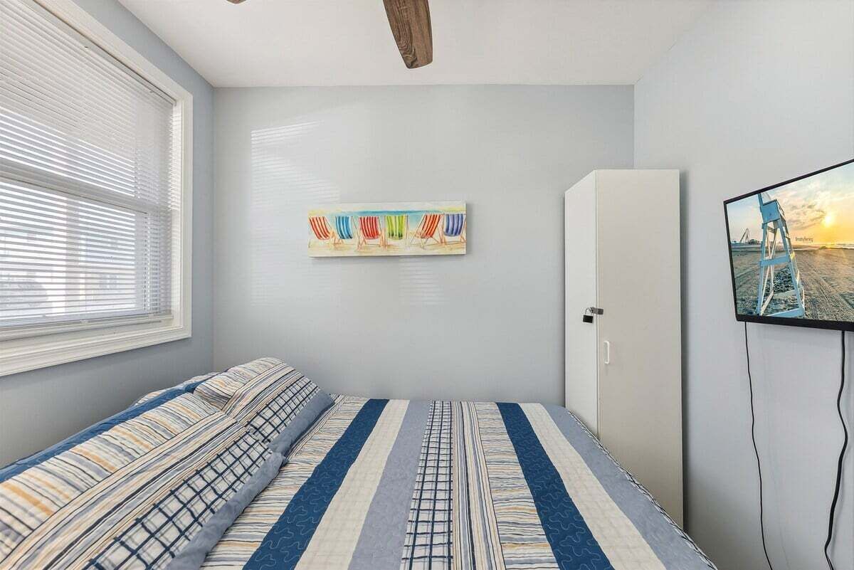 A bedroom with a bed, a window with blinds, a wall-mounted TV, and a cabinet.