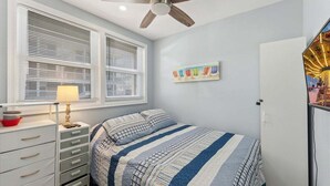 A bedroom with a bed, a ceiling fan, a window with blinds, a bedside table with a lamp, and a wall-mounted TV. - Modern 2 Bedroom Condo with Pool (Wildwood Crest)