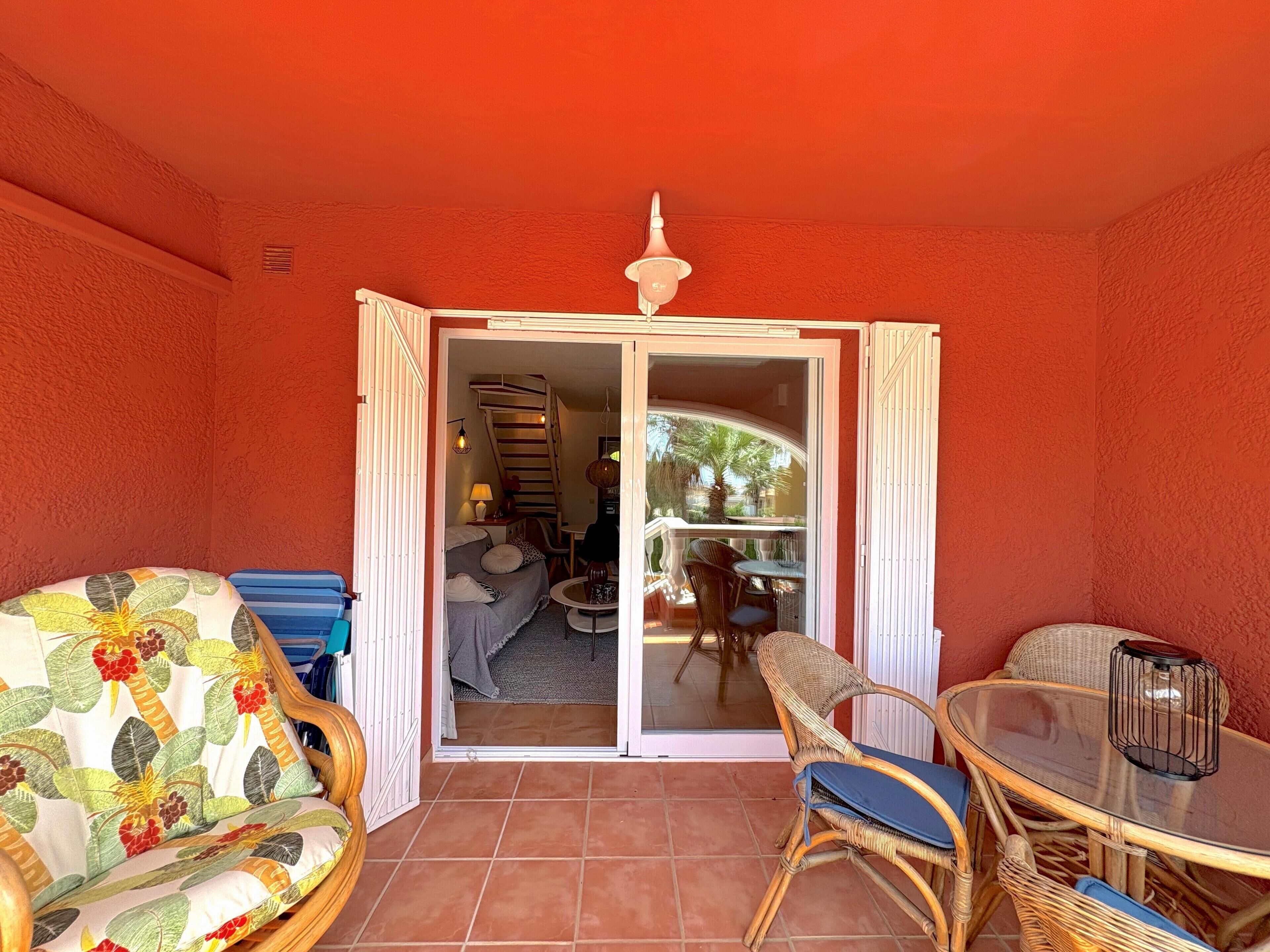 A room with a red wall, a sliding glass door leading to a balcony, wicker furniture, and a small table with a glass top.
