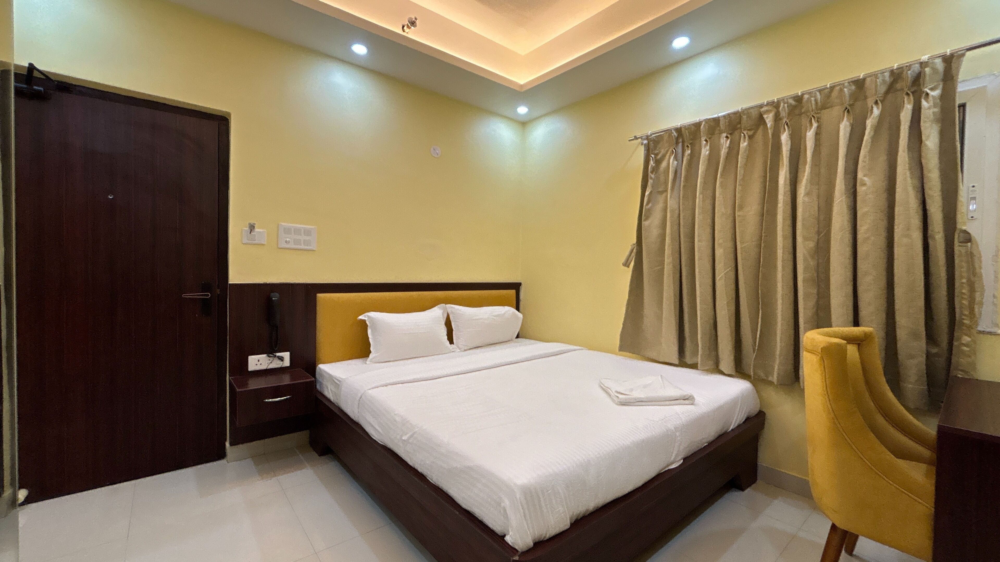 A hotel room with a large bed, a wooden headboard, a bedside table with a phone, a yellow armchair, and a curtain.