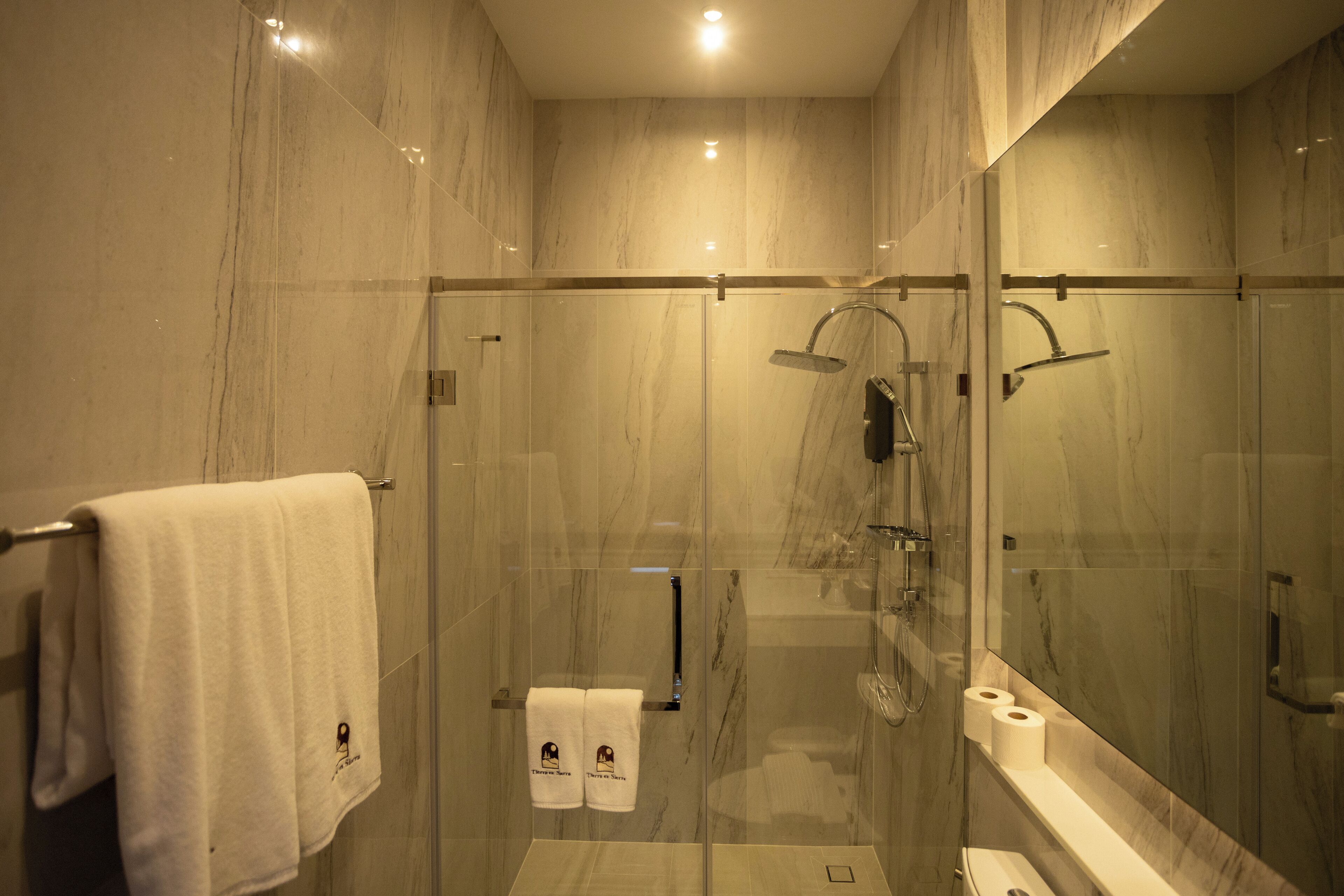 A modern bathroom with a glass-enclosed shower, marble walls, and a visible toilet paper holder.