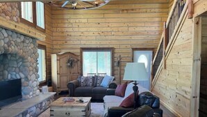 Interno di una baita di legno con camino in pietra, pareti in legno, una scala e un'accogliente area salotto.