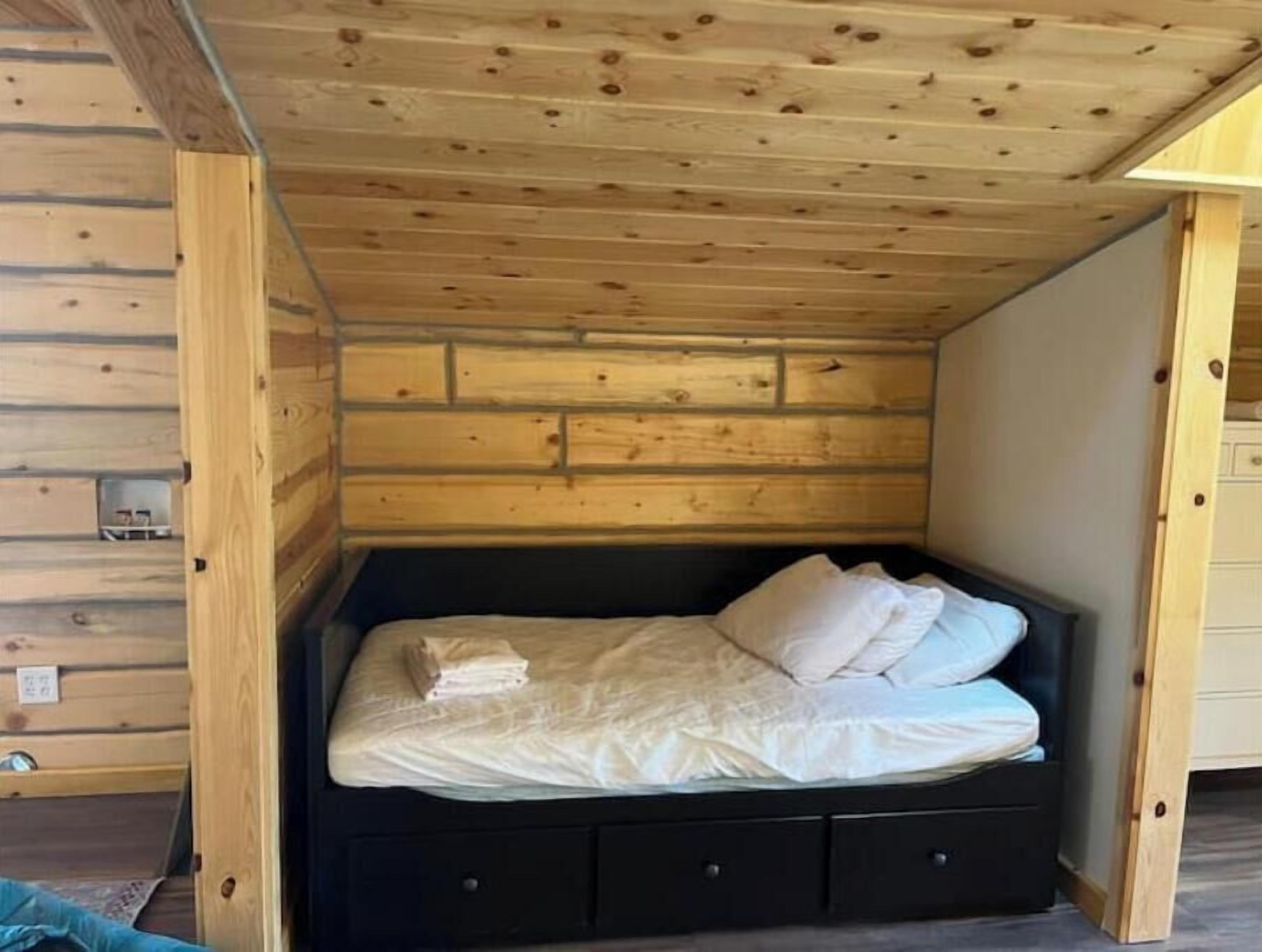 Une pièce de cabane en bois comprenant un lit, une petite commode et un plafond en bois.