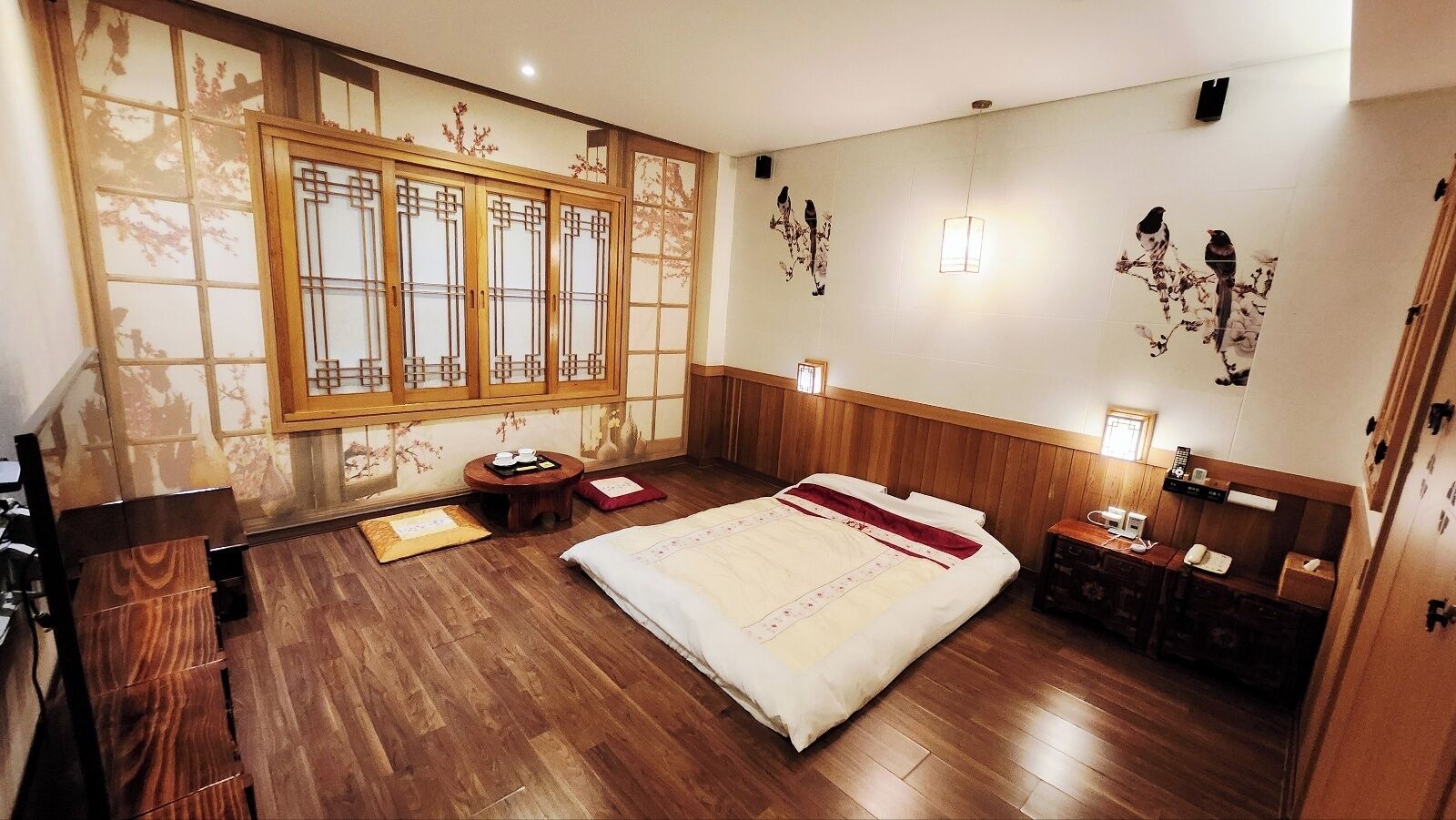 A traditional-style bedroom with a wooden floor, a large bed, a small round table, a nightstand, and a window with decorative paper.
