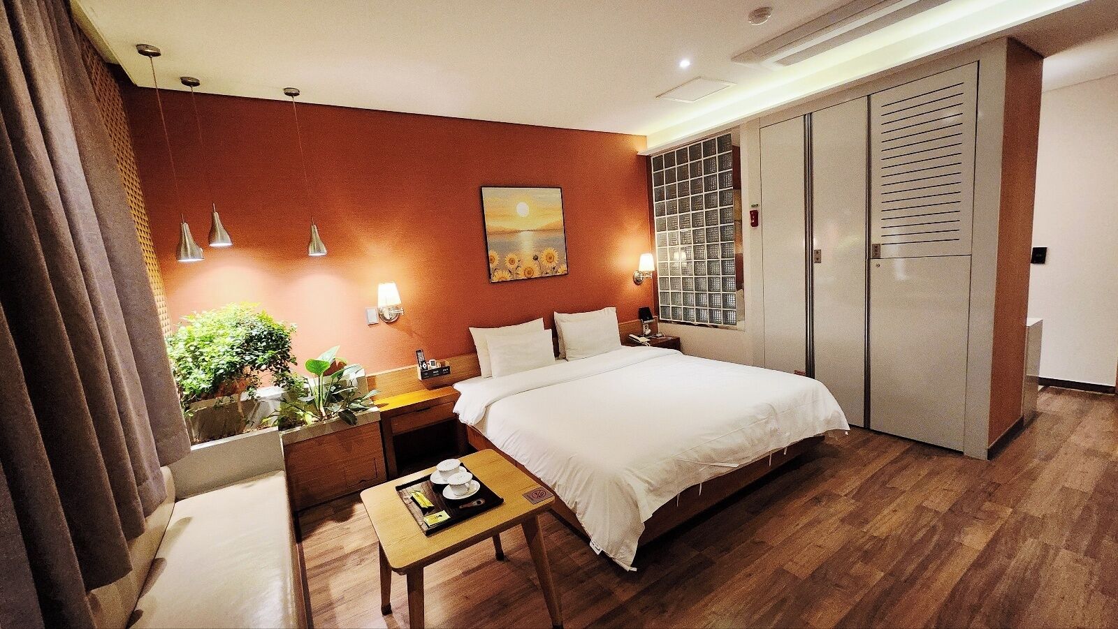 A hotel room with a bed, bedside table, lamp, and a view of the outdoors through a window.