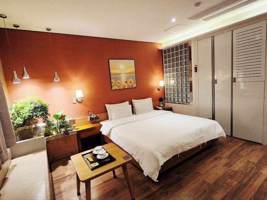 A hotel room with a bed, bedside table, lamp, and a view of the outdoors through a window.