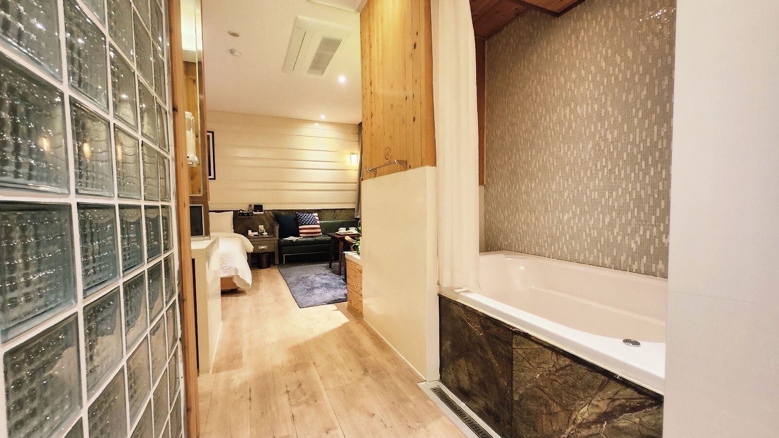 A narrow hallway leading to a bedroom with a bed, a small table, and a nightstand. There is a bathroom with a bathtub and a shower area.