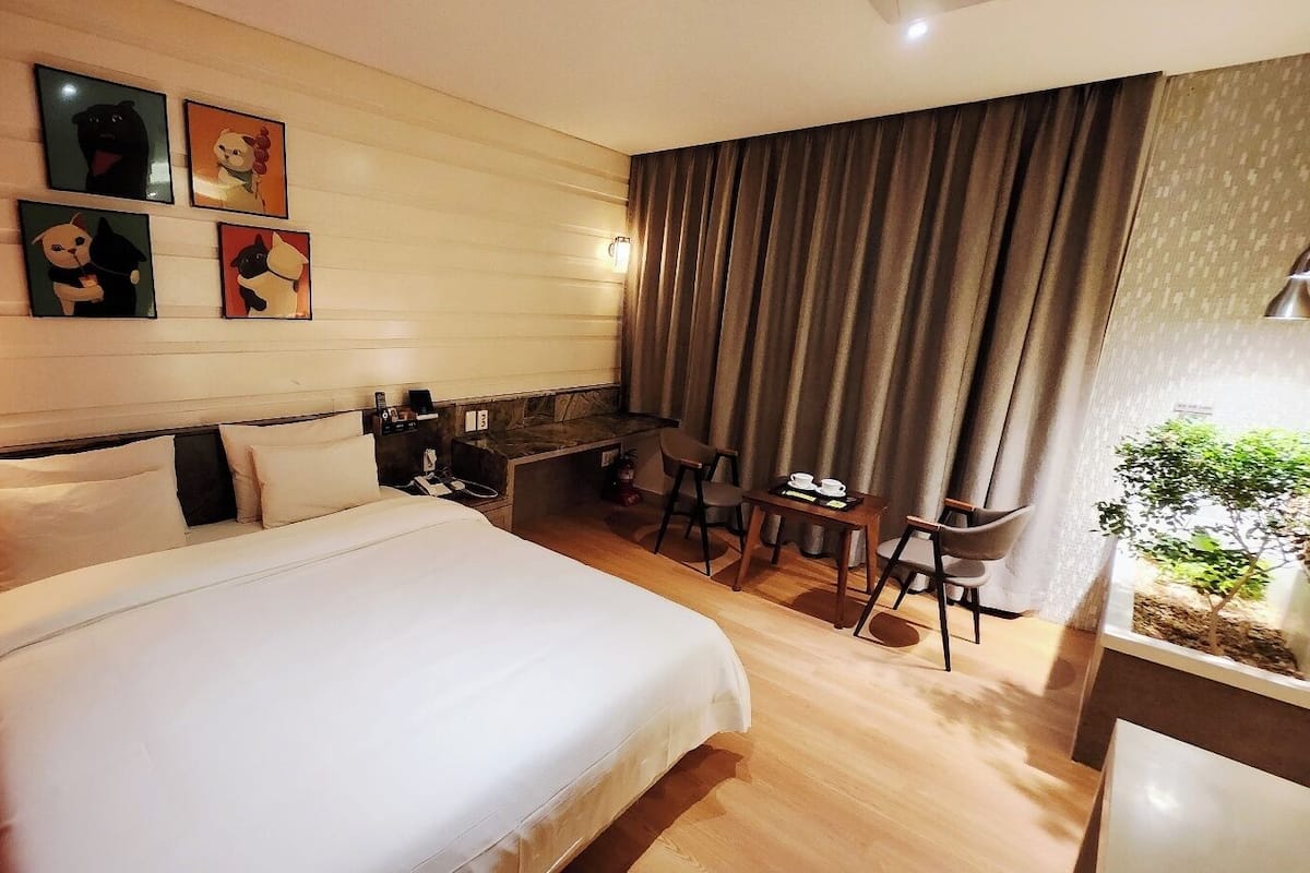 A hotel room with a large bed, a desk, and a chair. There are wall-mounted lights and a small plant on the windowsill.