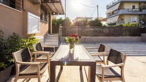 En udendørs patio med et træbord og stole, en vase med blomster, og udsigt til en boligbygning i baggrunden.