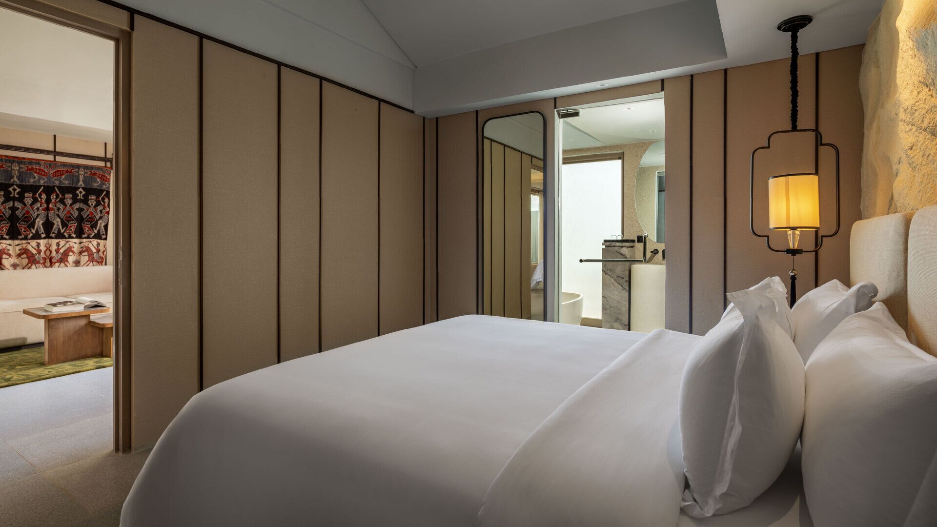 A hotel room with a large bed, a bedside lamp, a mirror, and a view of a bathroom and a balcony with a patterned curtain.