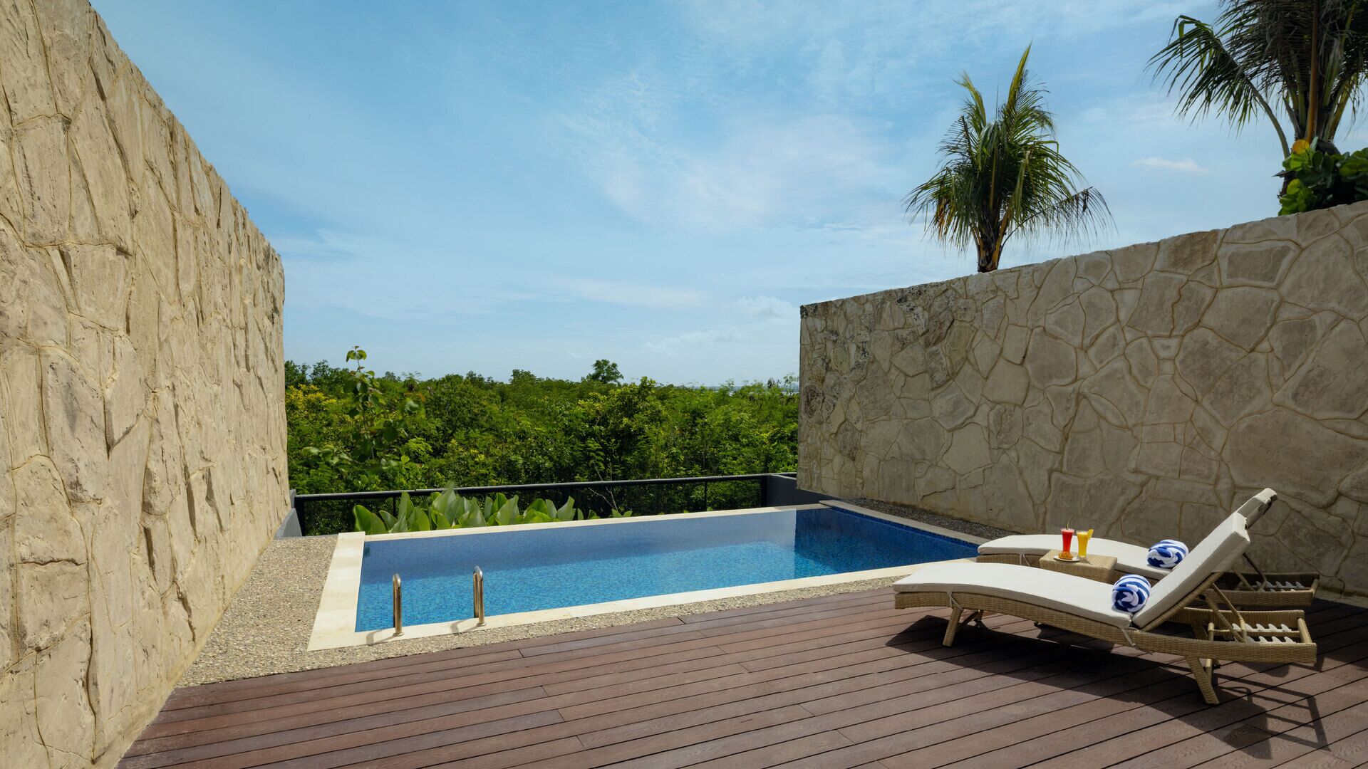 A pool area with a wooden deck, stone walls, and a lounge chair with sun loungers.