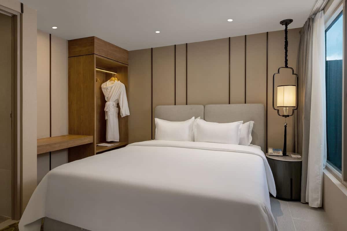 Habitación de hotel con una cama grande, una bata colgada en un estante de madera, una mesita de noche con lámpara y una ventana con cortinas.