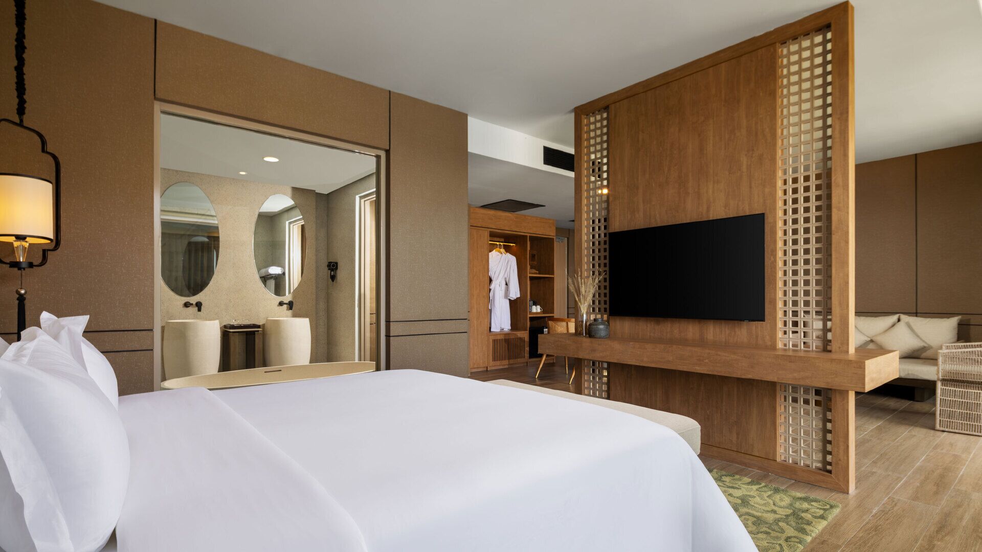 A hotel room with a large bed, a TV mounted on a wooden panel, a wardrobe, and a bathroom with a mirror and sink.
