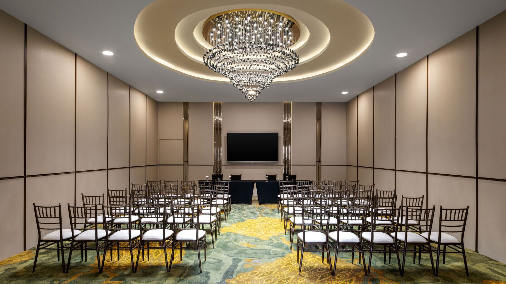 A conference room with a large chandelier, rows of chairs, a carpeted floor, and a flat-screen TV on the wall.