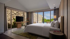 Una habitación de hotel moderna con una cama grande, un televisor de pantalla plana, una mesita con un libro y vistas a una piscina y vegetación.