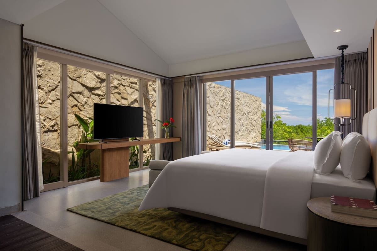 Una habitación de hotel moderna con una cama grande, un televisor de pantalla plana, una mesita con un libro y vistas a una piscina y vegetación.