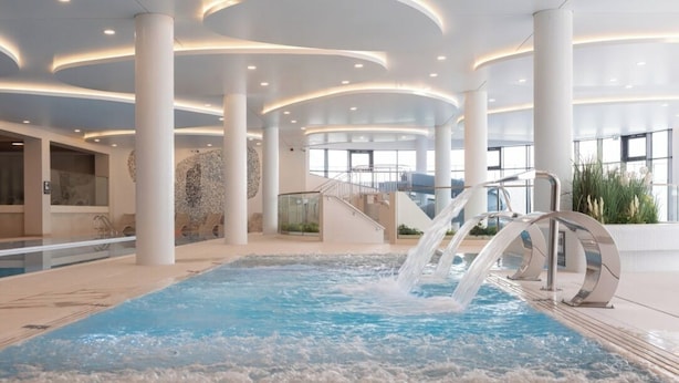 A modern indoor pool area with a central water feature, surrounded by white columns and glass railings.