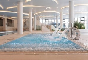 A modern indoor pool area with a central water feature, surrounded by white columns and glass railings. - Polanki Aqua Apartamenty - Family vacation with pool & wellness (Kolobrzeg)