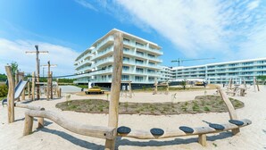 A playground with a seesaw, sand, and a building with multiple balconies in the background.