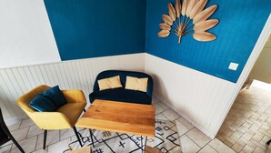 Quarto de canto com parede azul, cadeira amarela, sofá azul escuro, mesa de madeira e tapete estampado.