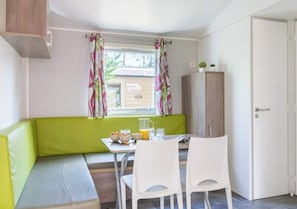 A small kitchenette with a window, a green cushioned bench, a table with chairs, and a cabinet. - Camping Le Ty Nadan (Locunolé)