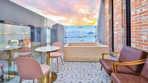 Un balcón con vista al mar, una bañera de madera, sillas rosas y una mesa redonda.
