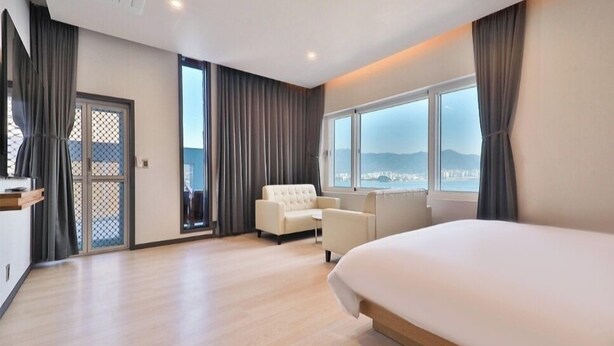 Una habitación de hotel moderna con una cama grande, una zona de estar con sofá y sillones, un ventanal con vistas a la ciudad y una puerta corredera que da a un espacio exterior.