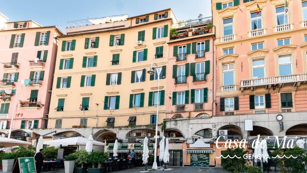 A row of multi-story buildings with balconies, a street in front with outdoor seating, and a sign for "Casa de Genova". - Casa Dè Mà, Large Luxury House with View of the Old Port and Aquarium of Genoa (Genova)