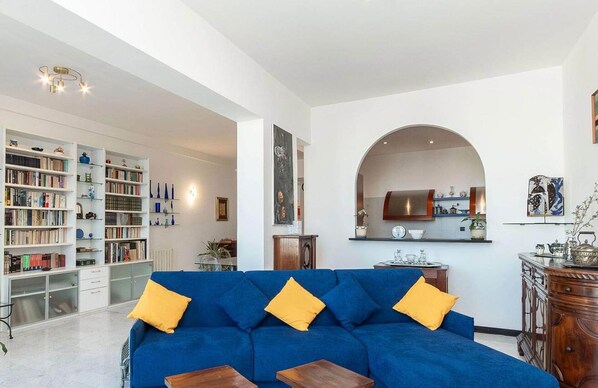 A living room with a blue sofa, yellow cushions, a bookshelf, and a kitchen visible through an archway. - Casa Dè Mà, Large Luxury House with View of the Old Port and Aquarium of Genoa (Genova)