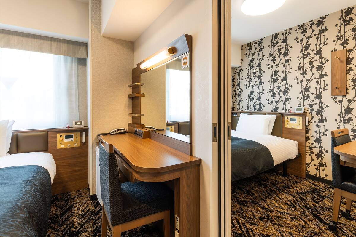 Ein Hotelzimmer mit Schreibtisch, zwei Betten, einem Spiegel, einem Nachttisch mit Telefon und einer Wand mit Blumenmuster.