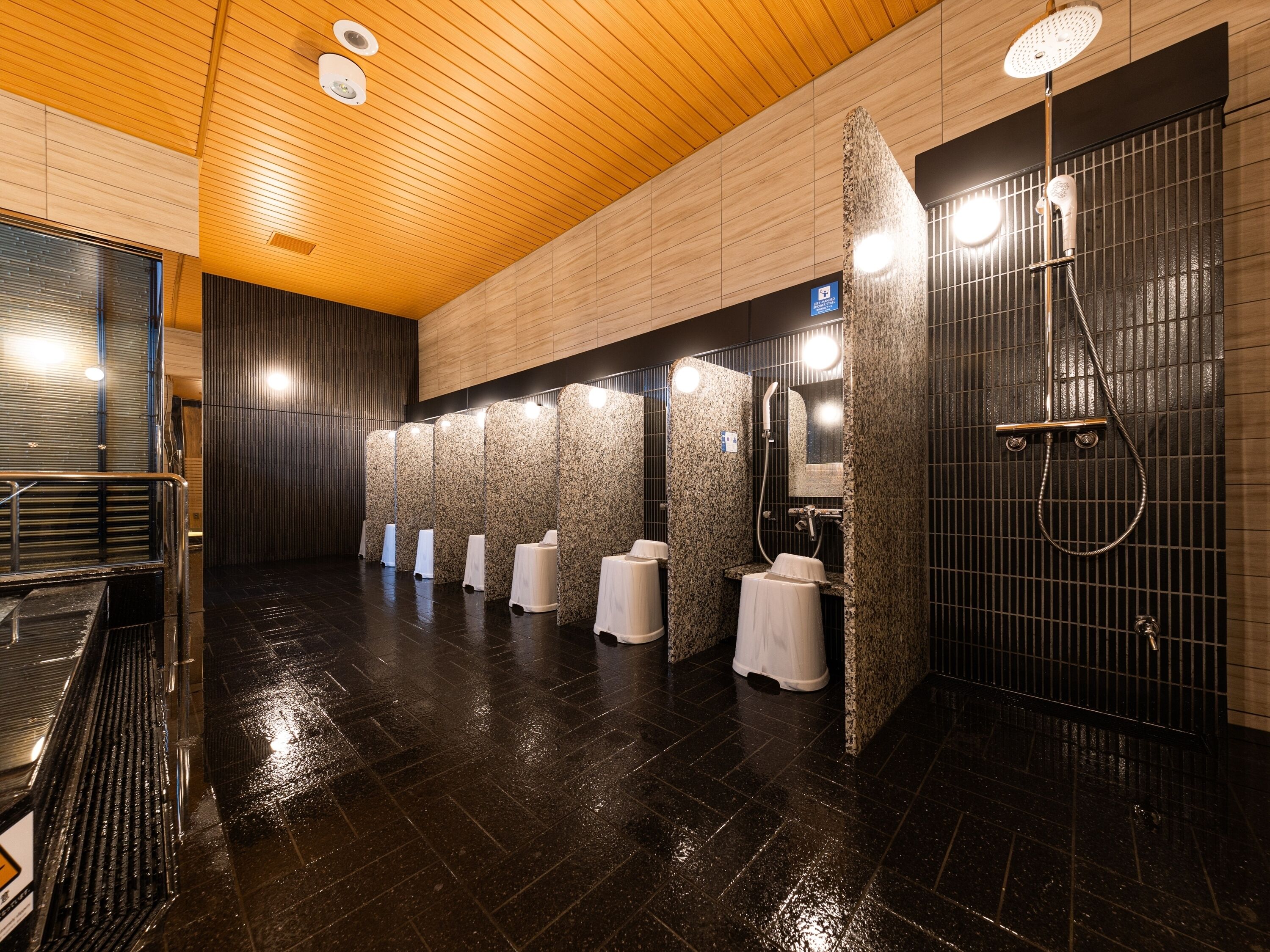A modern bathroom with a wet floor sign, a wooden ceiling, and a black tiled floor.