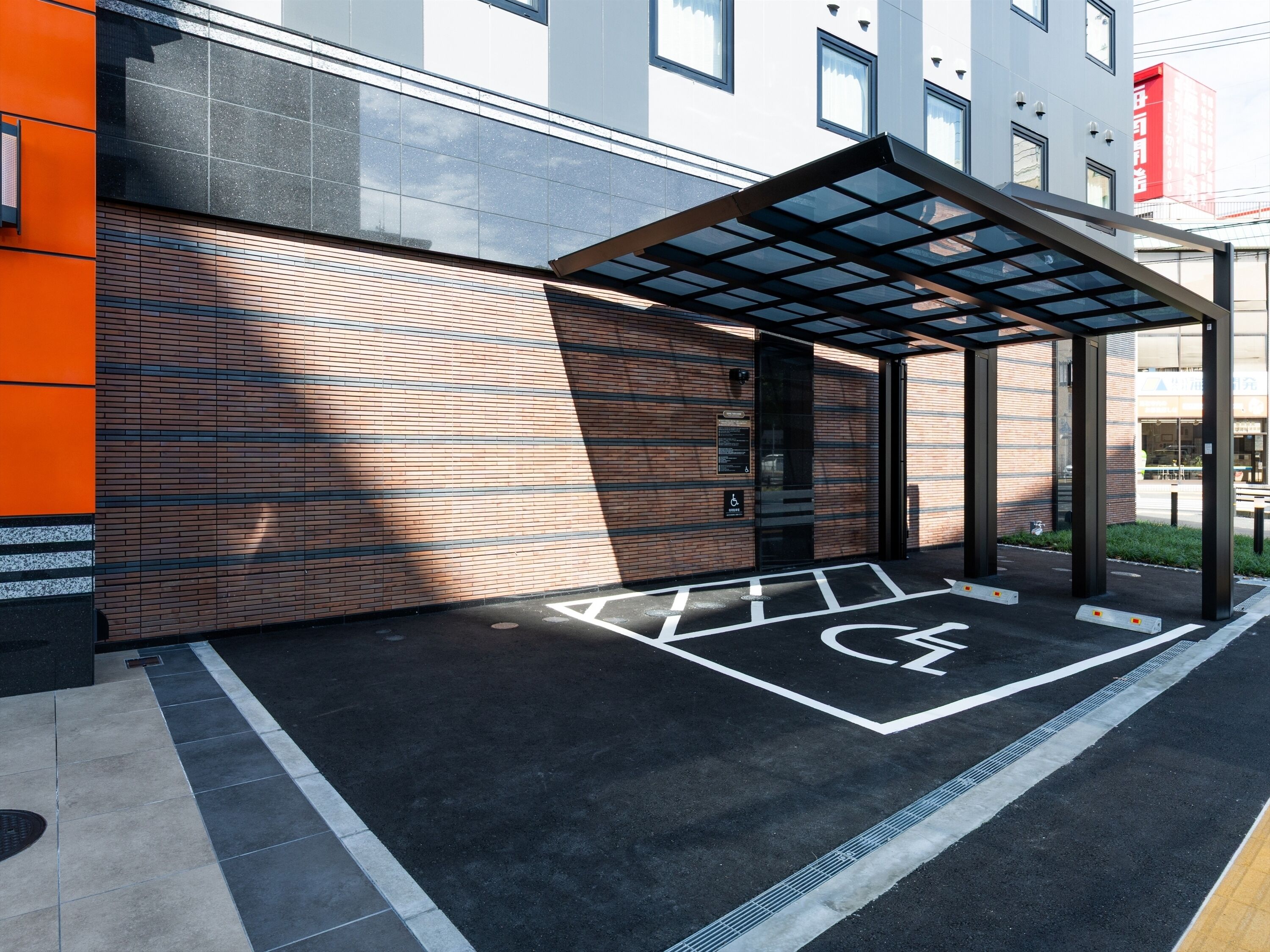 A modern building entrance with a covered walkway, a wheelchair-accessible ramp, and a parking space marked with accessibility symbols.