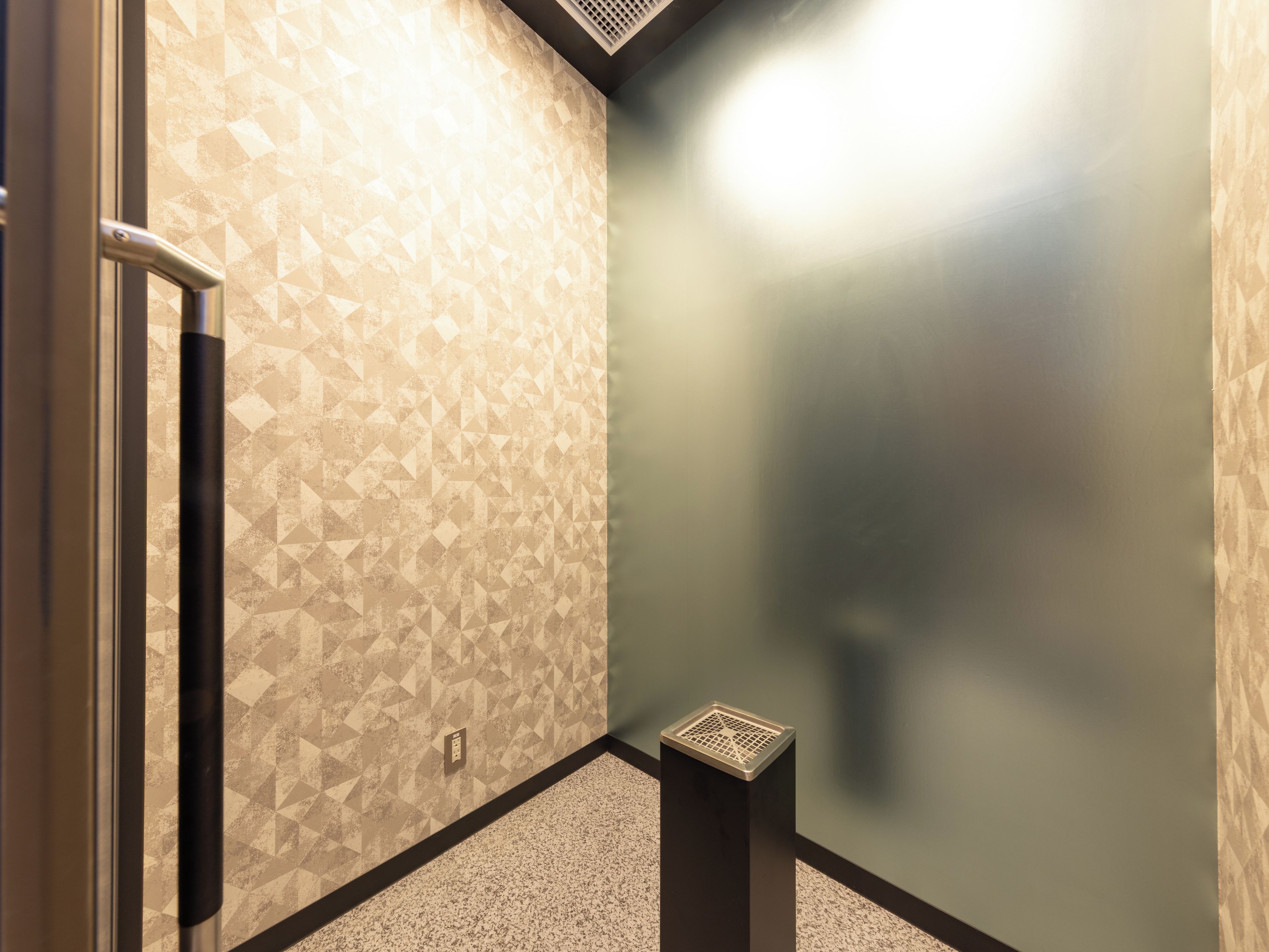 A corner of a room with patterned walls, a door, and a small trash bin.
