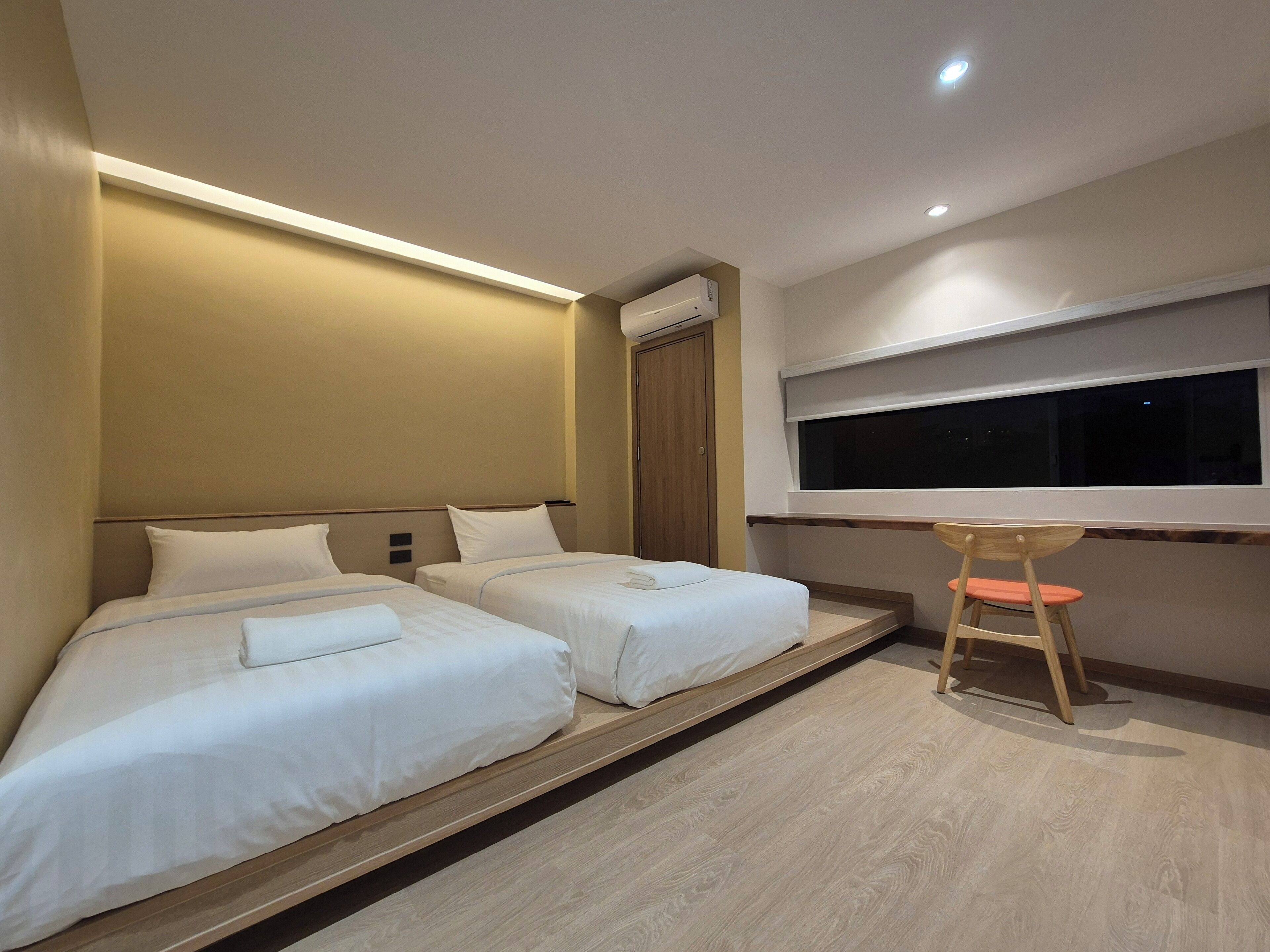 A hotel room with two beds, a small desk with a chair, a window, and a ceiling light.