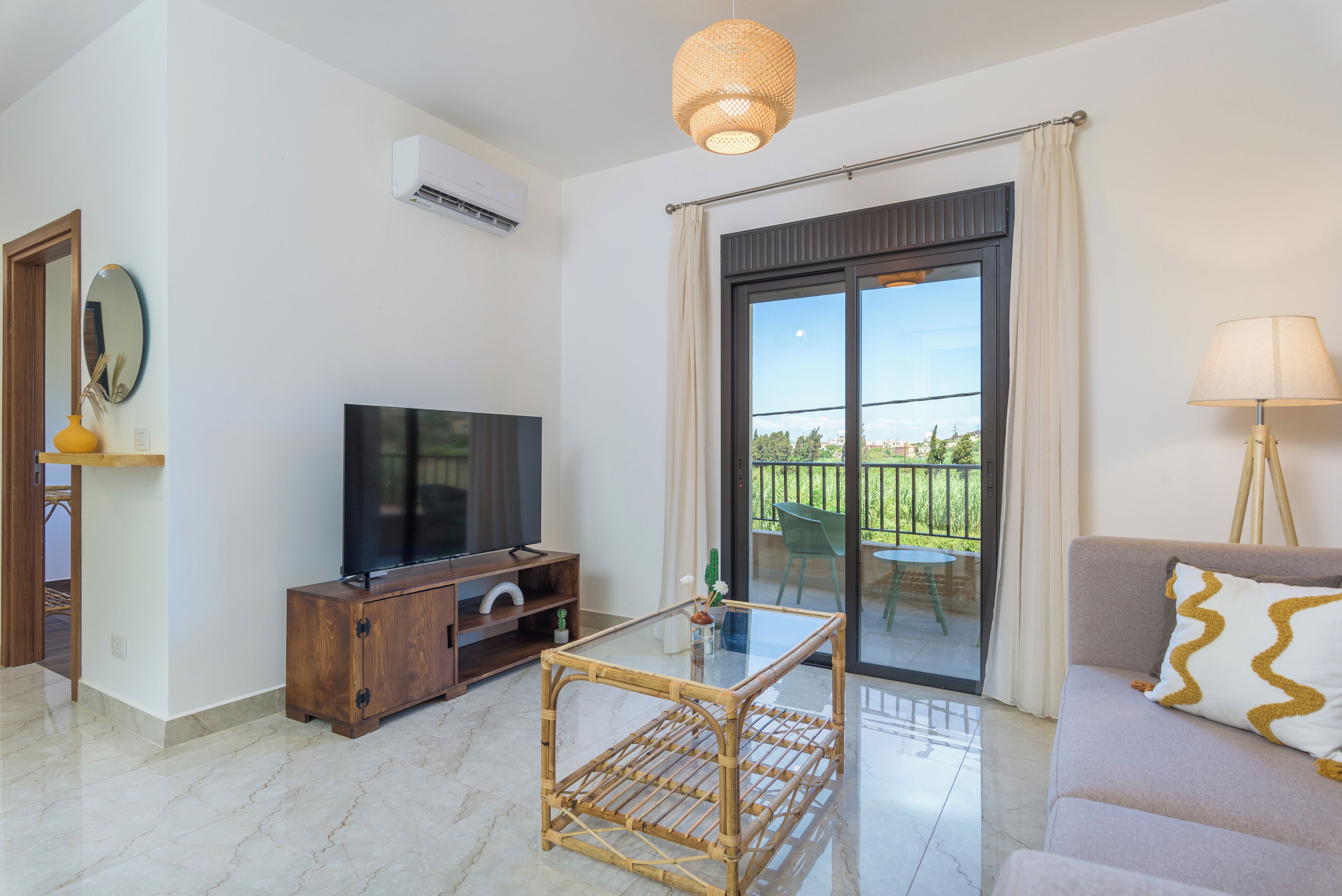 A modern living room with a flat-screen TV, a glass-top coffee table, a sofa, and a balcony with a view.