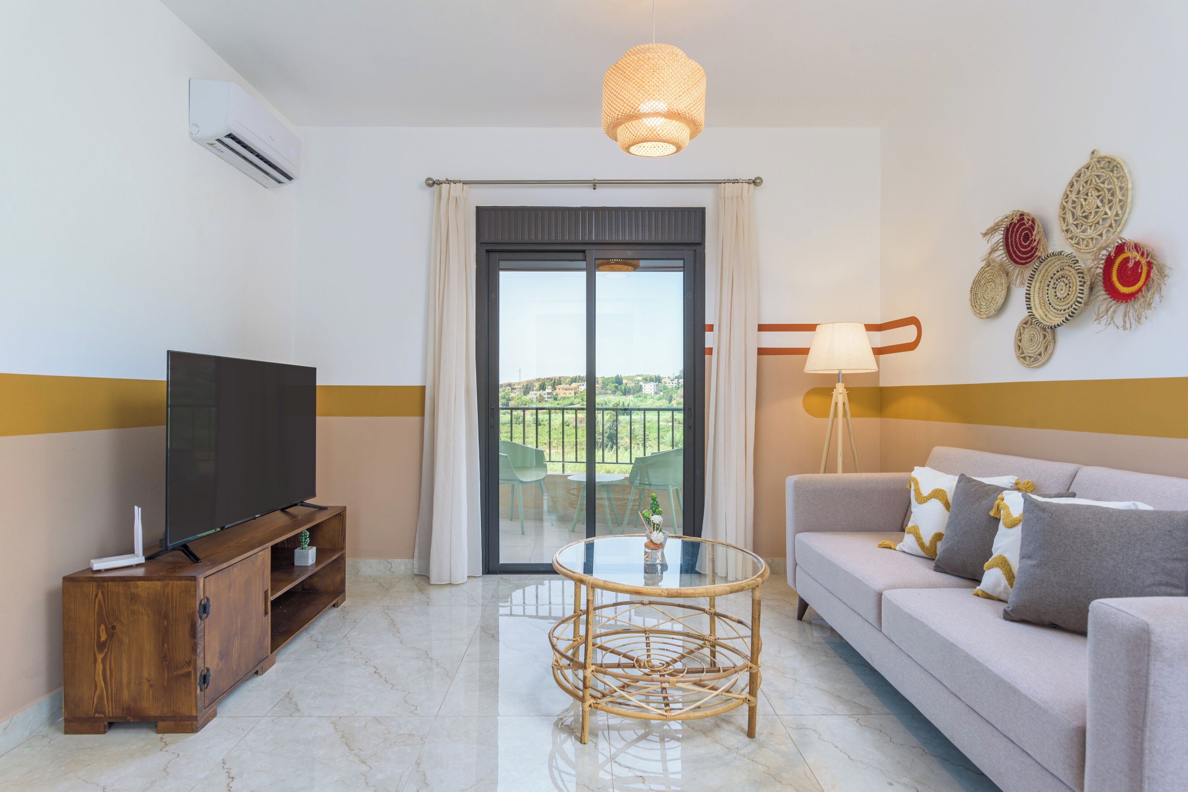 A modern living room with a flat-screen TV, a glass-top coffee table, a sofa with patterned cushions, a floor lamp, and a balcony with a view.