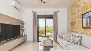 A modern living room with a flat-screen TV, a sofa, a coffee table, and a view of a garden through large windows.