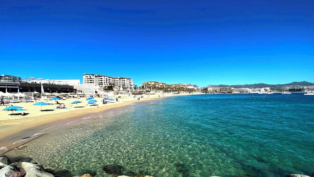 A beach with clear water, a sandy shore, and a hotel in the background. - Casa M Cabo, Modern Luxury Villa, Private Pool & Hot Tub (Cabo San Lucas)