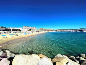 A beach with clear water, a sandy shore, and a hotel in the background.