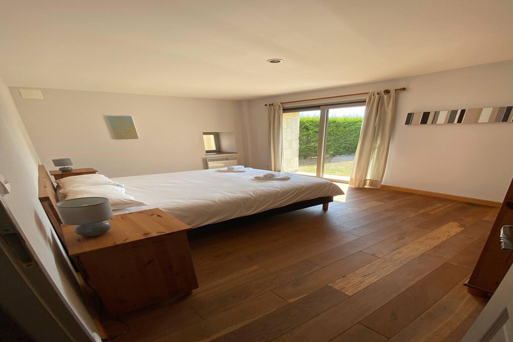 A bedroom with a large bed, a bedside table, a nightstand, a window with curtains, and a view of greenery outside.