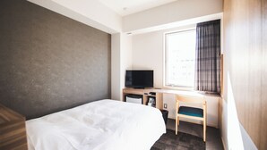 Ein Hotelzimmer mit einem Bett, einem Schreibtisch mit Fernseher, einem Fenster mit Vorhängen und einem kleinen Stuhl.